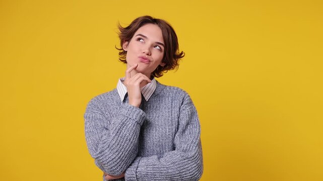 Young woman wear grey knitted sweater white shirt casual clothes look around think dream put hand prop up on chin lost in thought and conjectures isolated on plain yellow background. Lifestyle concept