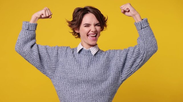 Strong sporty fitness young woman wear grey knitted sweater white shirt casual clothes show biceps muscles on hand demonstrating strength power isolated on plain yellow background. Lifestyle concept
