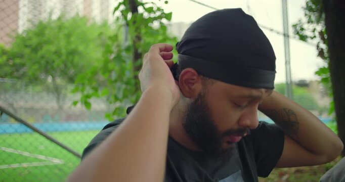 African American man tying and adjusting black durag outdoors near park fence, showing focus, care, and pride in personal style and cultural expression in everyday urban life