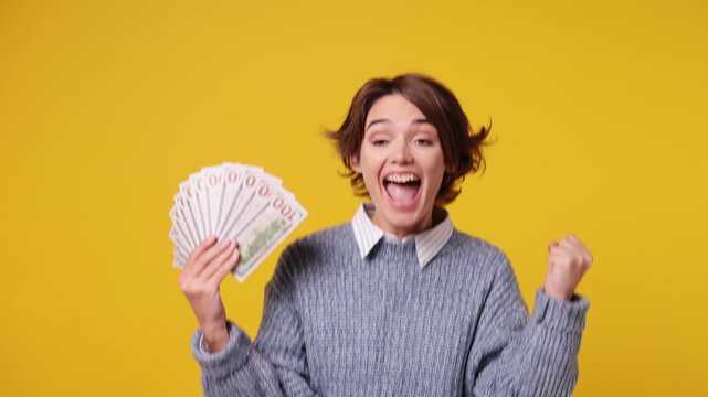 Happy young woman wears grey knitted sweater white shirt casual clothes hold fan of cash money in dollar banknotes do winner gesture celebrate isolated on plain yellow background. Lifestyle concept