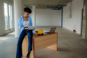 Young female architect sitting on a desk using a digital tablet while inspecting the progress of a construction site in a modern, empty office space