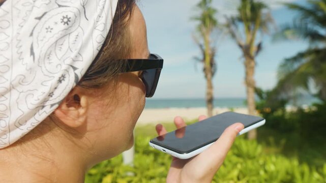 Woman recording voice message on smart phone. Using virtual assistant for communication. Hands free. Enjoying tropical beach vacation. Wearing sunglasses and bandana. Modern mobile technology concept