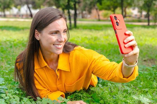 Happy woman wearing yellow shirt lying on green grass outdoors taking selfie with smartphone, concept of leisure, social media, connection, positivity, nature, lifestyle, relaxation. - Powered by Adobe