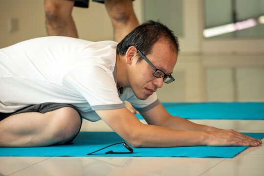 Middle-aged Asian man practicing yoga in child's pose on a blue mat indoors. He appears focused and calm, representing mindfulness, flexibility, and wellness for seniors or beginners in yoga practice