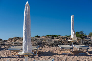 Closed umbrellas and empty sun loungers on a quiet sandy beach under a clear blue sky.