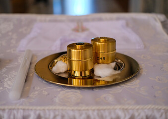 Golden chrism containers labeled “CHR” placed on a silver tray with cotton on an altar cloth in a church.