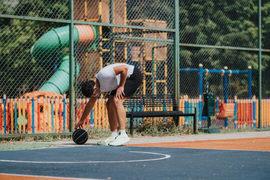 A man in a white shirt and shorts retrieves a basketball on an outdoor court, with a colorful playground and fence in the background, capturing focus, fitness, and active recreation.