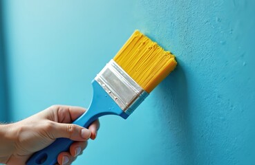 Hand holding a blue paint brush with yellow bristles applies paint to blue wall surface. Brush is poised to apply paint. Renovation or home improvement concept.