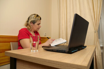 A woman works at a laptop, occasionally taking notes in a notebook.