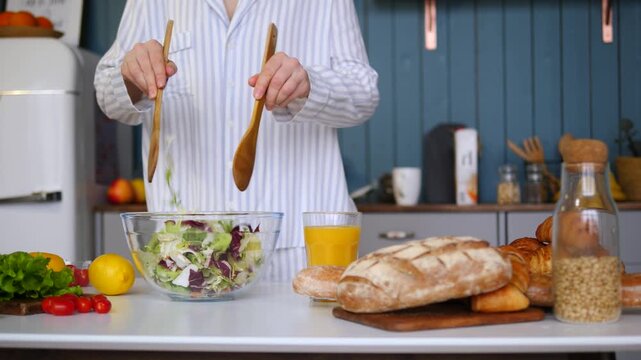 Young woman in pajamas preparing a healthy breakfast in the kitchen, tossing a fresh green salad with wooden servers. A nutritious meal to start the day, promoting wellness and a balanced lifestyle