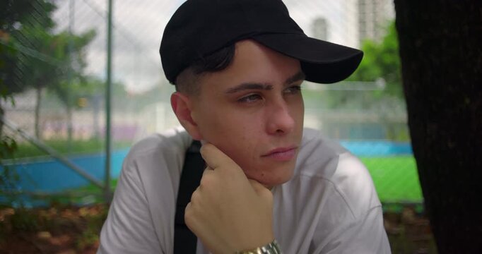 Young Latin man sitting outdoors near soccer field, looking away thoughtfully with calm expression, wearing black cap and casual clothes, evoking reflection and contemplation