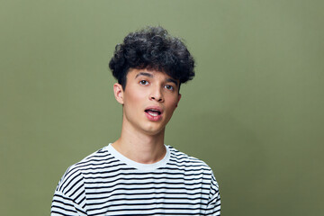 Young man with curly hair wearing a striped shirt against a muted green background, expressing...