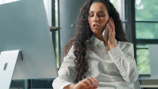 Smiling Hispanic business woman talking phone looking computer businesswoman female girl employer multitasking in office work professional with smartphone consulting client talk typing pc internet