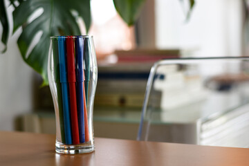 Several colorful marker pens stand upright in a clear drinking glass on a wooden table.