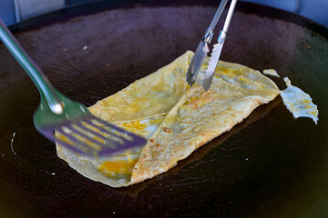 An egg crepe is cooked on a hot griddle, being folded with a spatula and tongs.