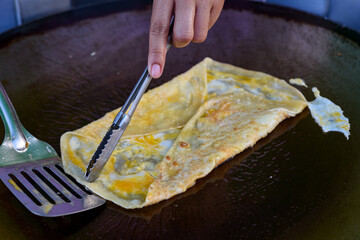An egg crepe is cooked on a hot griddle, being folded with a spatula and tongs.