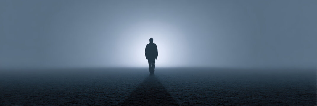 Silhouette of man walking alone into dark fog toward mysterious light. lonely, conceptual journey into an unknown future filled with mystery and emotion