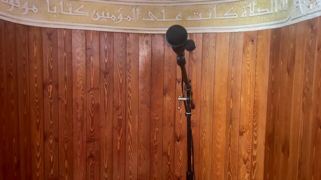 A peaceful wooden prayer corner inside an empty mosque, with a microphone stand prepared for the imam&rsquo;s sermon, soft patterned carpet, serene lighting capturing the calm atmosphere of Islamic worship