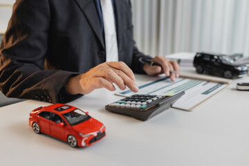A businessman in a suit is calculating car loan options using a calculator and comparison chart, with model cars and car keys on the desk, symbolizing finance, leasing, and investment planning.