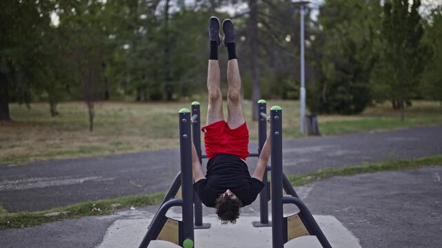 Determined strong man performing an intense core workout, hanging upside down from parallel bars in a public park. He demonstrates strength and dedication to fitness and health.