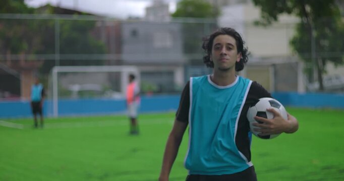 Hispanic man holding soccer ball standing on turf field, looking confidently at camera while teammates play in background, showing pride and focus during practice