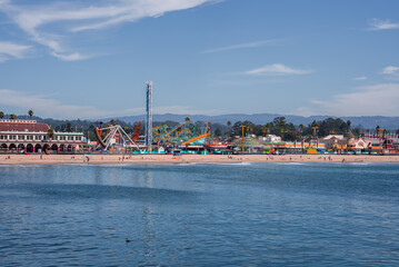 Wide daytime panorama shows the Santa Cruz Beach Boardwalk in California, the colonnaded Casino Arcade at left, rides along the sand, Monterey Bay water, and visitors on the shore.