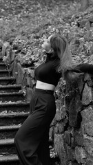 A woman in a black crop top and wide-leg pants stands by a stone wall on the steps in a park. She has her head thrown back and is relaxedly breathing in the fresh air, enjoying a moment of peace.