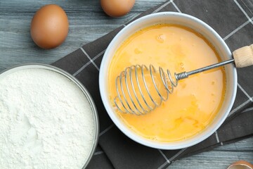 Beaten eggs in bowl, whisk and flour on wooden table, flat lay