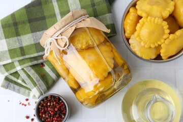 Pickled pattypan squashes in jar, bowl, oil and peppercorns on white tiled table, flat lay