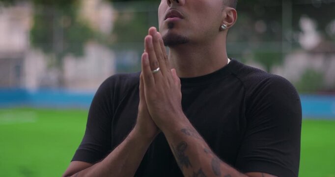 Hispanic man of African descent standing outdoors with hands clasped in front of face, pensive and slightly worried expression, looking up with hope and quiet faith in moment of reflection