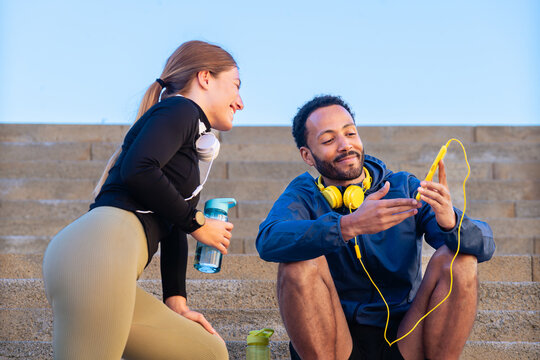 Athletic man and woman resting on stairs after training, smiling and sharing music from smartphone. Sporty friends with headphones and water bottles enjoying outdoor fitness break and motivation