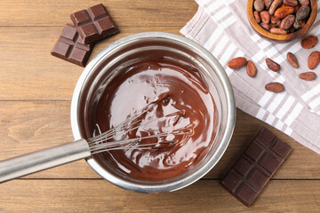 Melted chocolate with whisk in bowl, beans and pieces on wooden table, flat lay