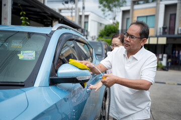 Middle aged Asian couple enjoys quality time together washing their blue car on a sunny day