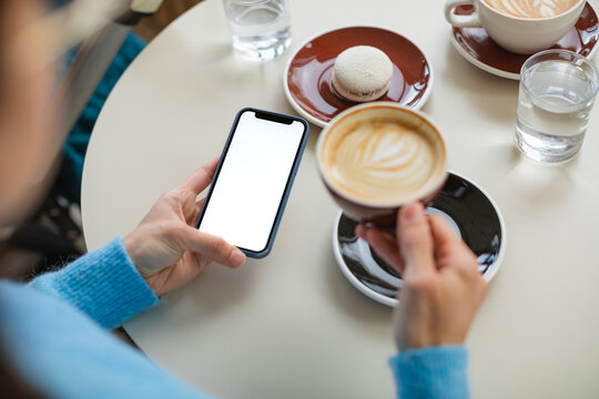 Overhead view of a woman sitting in a cafe with a cappuccino and a vanilla and coconut macaron holding a mobile phone with a blank screen