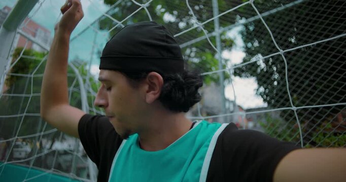 Hispanic soccer player holds goal net in thoughtful silence after match, looking away as he reflects on life, choices, and personal meaning on quiet urban field