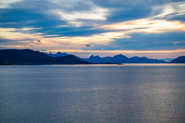 A serene seascape at dusk features a vast body of water with distant mountains under a sky painted with soft hues of blue and orange, Sortland, Norway