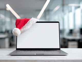 Open laptop with blank white screen and red Santa hat placed on top corner. Modern office background with soft daylight and Christmas atmosphere. Copy space on screen for festive design or text