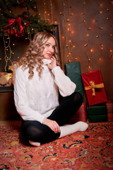 A beautiful girl was sitting on the floor, surrounded by gifts that were presented to her in honor of the holiday. She is wearing a white sweater and dark trousers. The trend of Christmas celebrations