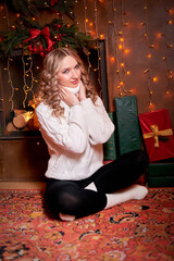 A beautiful girl was sitting on the floor, surrounded by gifts that were presented to her in honor of the holiday. She is wearing a white sweater and dark trousers. The trend of Christmas celebrations
