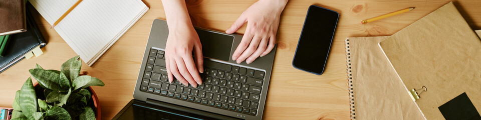 Website banner of hands of woman typing on laptop at wooden desk, smartphone and notebooks surrounding workspace