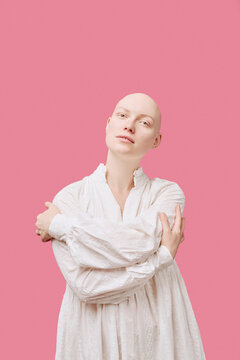 Portrait of young adult Caucasian woman with alopecia standing against pink background, crossing arms over chest, looking directly at camera with neutral expression