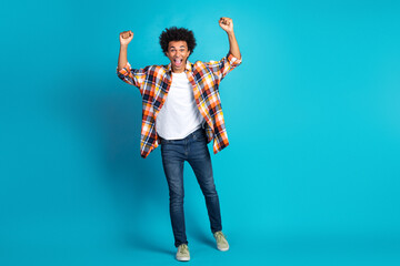 Young man celebrates with arms raised in casual plaid shirt and jeans against blue background expressing joy energy and confidence