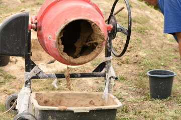 Freshly mixed clay is being poured from a red cement mixer into a container on green grass. One bucket is placed nearby. Outdoor construction or DIY work scene.