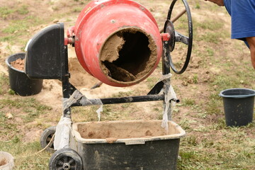 Freshly mixed clay is being poured from a red cement mixer into a container on green grass. Two black buckets are placed nearby. Outdoor construction or DIY work scene.
