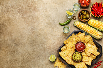 Nachos corn chips with guacamole and salsa dressing, fresh corn cobs, marinated jalapeño, chili peppers and tequila shots on beige concrete background. Mexican party snack. Top view with copy space