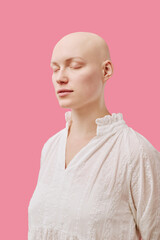 Caucasian young adult woman with alopecia standing with eyes closed against pink background, showing smooth bald scalp and serene facial expression, wearing light textured blouse