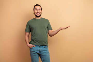 Young man in casual olive green t shirt poses with open hand against beige background smiling for lifestyle advertising and fashion shoot