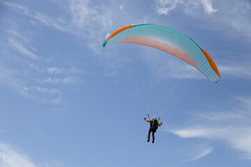 A paraglider with a vibrant blue and orange wing soars high above in a clear blue sky dotted with clouds.