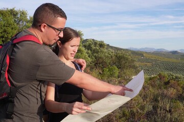 A man points at a map while explaining the route to a woman during a summer hike, surrounded by olive trees and a sunny rural landscape.