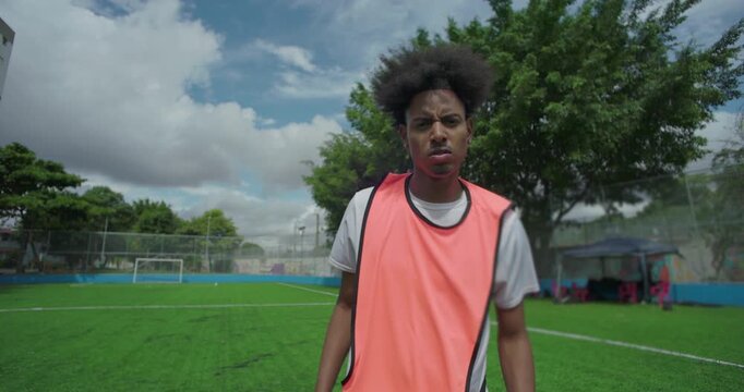 Young man in orange training bib on soccer field, serious and determined expression, afro hairstyle, showing focus and determination in urban sports environment